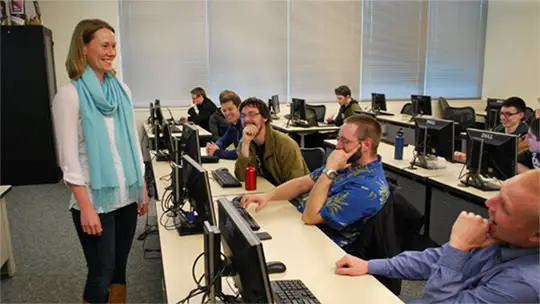 Students in a computer lab classroom paying attention to the instructor at the front of the classroom