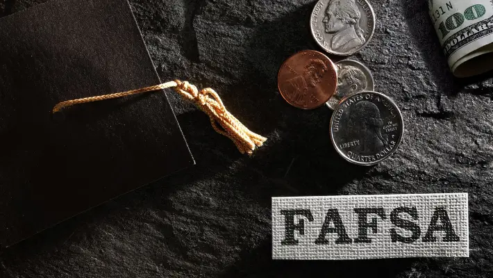 A graduation cap, money and a small cross stitched FASFA acronym laid out upon a table. 