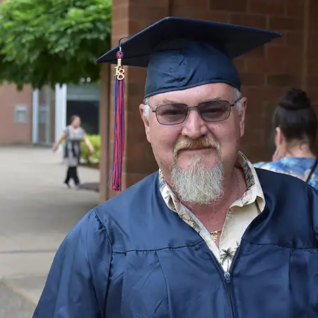 Older student smiles in cap and gown with a collared palm tree print shirt visible underneath