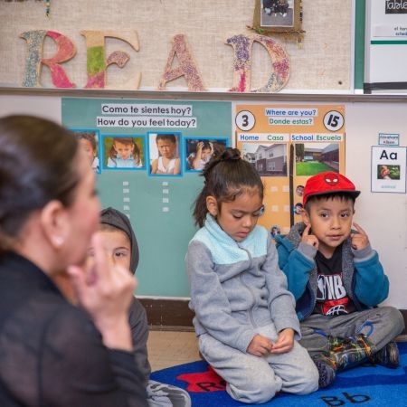 A teacher sits on the floor with three young children in a classroom. The walls display colorful educational posters.