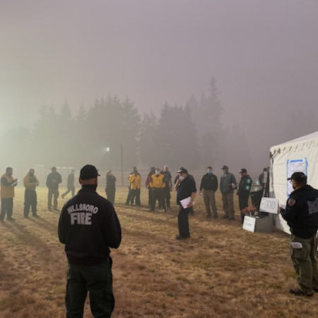 A diverse group of emergency management personnel — ranging from fire fighters to EMTs to incident managers — standing in an open field during wildfires making emergency coordination plans.