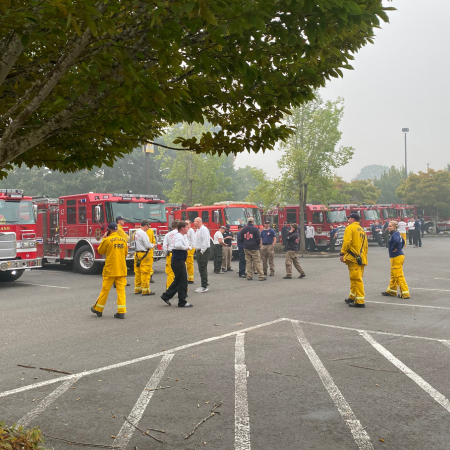 A diverse group of emergency management personnel — ranging from firefighters to EMTs to incident managers — standing in a parking lot during wildfires making emergency coordination plans.
