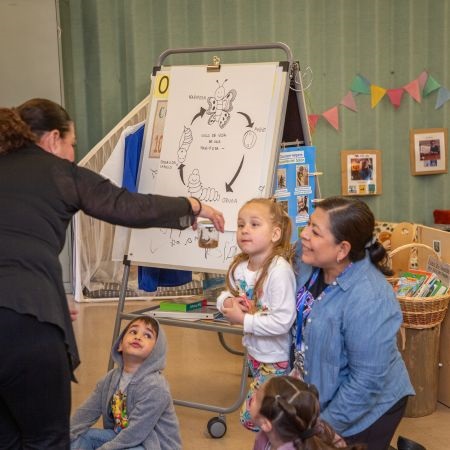 A teacher shows an attentive child a small jar with a caterpillar. Behind them is a diagram on an easel about the butterfly lifecycle.
