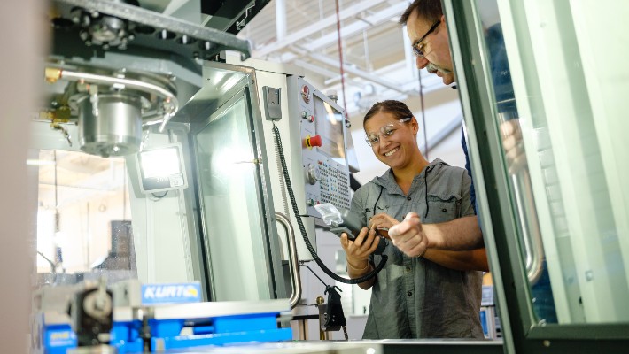 A machine tool student smiling while being instructed on how to use the machine in front of her.